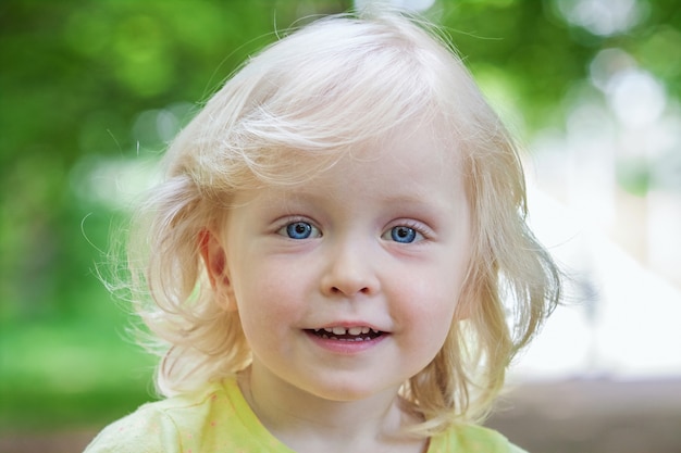 Linda niña en edad preescolar de pelo rubio con ojos azules sonríe alegremente en el fondo de la vegetación en el parque