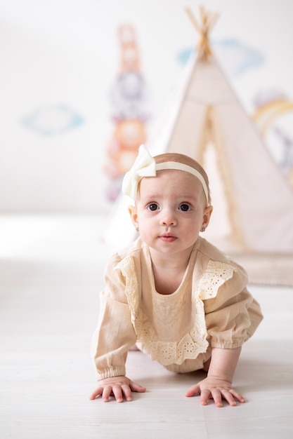 Una linda niña europea con ojos marrones en un traje beige hecho de tela natural juega en la habitación de los niños en casa con el telón de fondo de un wigwam y juguetes de peluche
