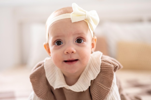 Una linda niña europea con ojos marrones en un traje de punto beige está sentada en la cama en casa sonriendo un retrato de cerca de un niño