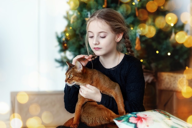 Una linda niña feliz abraza a un gato abisinio en el fondo de un árbol de Año Nuevo Hay luces