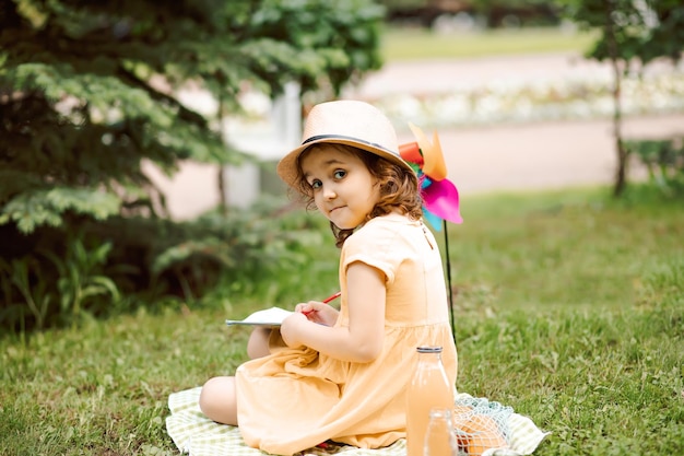 Linda niña feliz en un sombrero divirtiéndose en el prado del parque Niño sentado en una manta dibujando y mirando a la cámara Picnic en la naturaleza