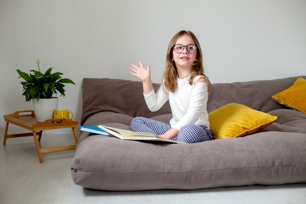 Linda niña con gafas, jeans y un cuello alto blanco está leyendo un libro sentado en la cama