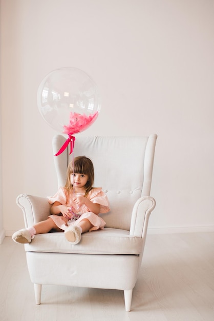 Linda niña con globo con elegante vestido rosa sentado en un sillón