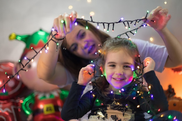 Linda niña haciendo payasadas con luces de Navidad, su madre la mira con asombro