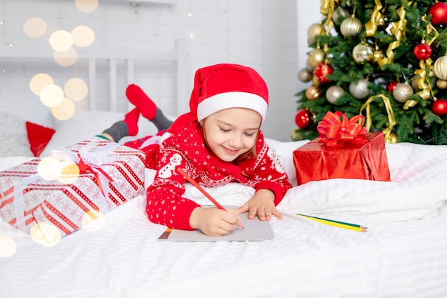 Linda niña hermosa escribe una carta a Santa Claus en el árbol de Navidad con un suéter rojo y un sombrero en la víspera de Año Nuevo o Navidad en casa en una cama blanca