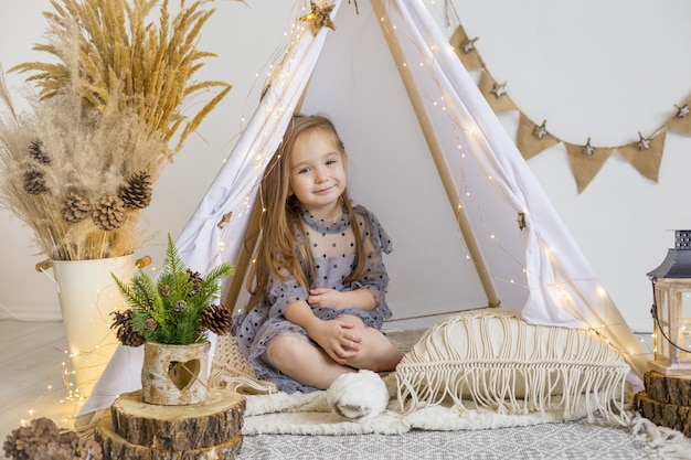 Una linda niña con un hermoso vestido juega en un wigwam en casa. Decoración de año nuevo.