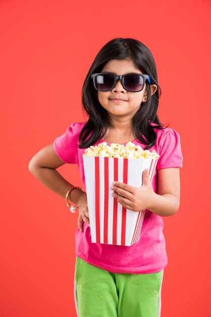 Linda niña india comiendo palomitas de maíz mientras usa gafas de sol o gafas 3D en un teatro. Se encuentran aisladas sobre fondo de colores