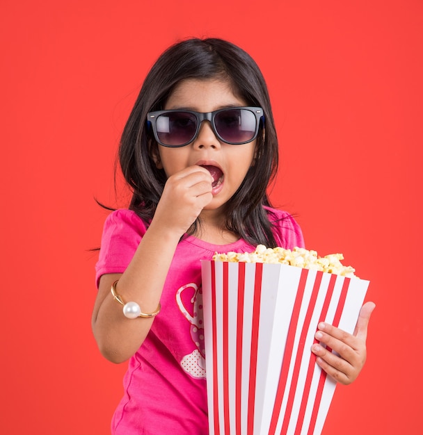 Linda niña india comiendo palomitas de maíz mientras usa gafas de sol o gafas 3D en un teatro. Se encuentran aisladas sobre fondo de colores