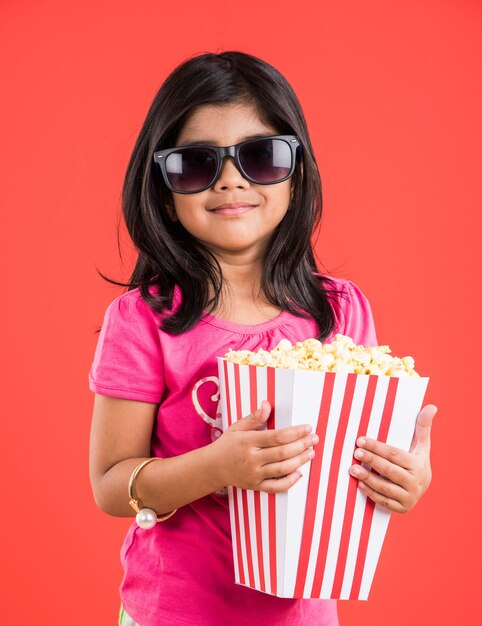 Linda niña india comiendo palomitas de maíz mientras usa gafas de sol o gafas 3D en un teatro. Se encuentran aisladas sobre fondo de colores