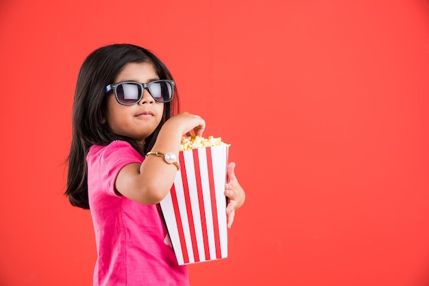 Foto linda niña india comiendo palomitas de maíz mientras usa gafas de sol o gafas 3d en un teatro. se encuentran aisladas sobre fondo de colores