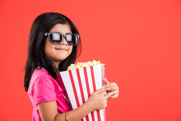 Linda niña india comiendo palomitas de maíz mientras usa gafas de sol o gafas 3D en un teatro. Se encuentran aisladas sobre fondo de colores