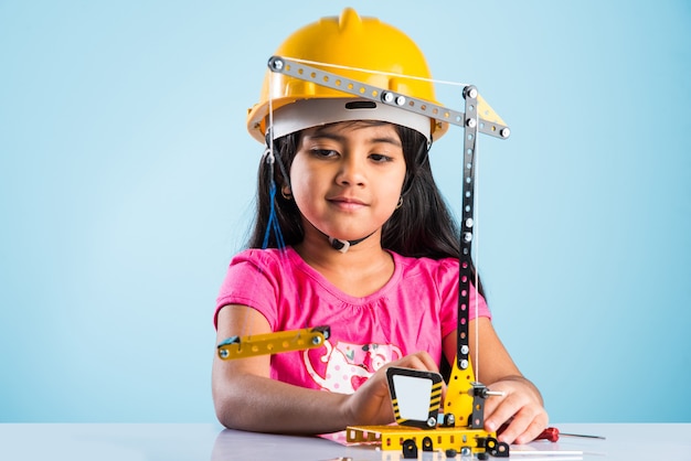 Linda niña india jugando con grúa de juguete con sombrero de construcción amarillo o casco, concepto de infancia y educación