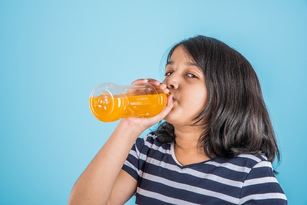 Linda niña india o asiática con una botella de bebida fría de naranja o mango o jugo de frutas, bebiendo o sosteniendo mientras está de pie aislado sobre fondo azul o blanco.