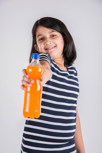 Linda niña india o asiática con una botella para mascotas de bebida fría de naranja o mango o jugo de frutas, bebiendo o sosteniendo mientras está de pie aislado sobre fondo azul o blanco.