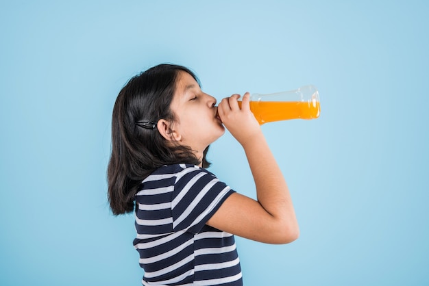 Linda niña india o asiática con una botella para mascotas de bebida fría de naranja o mango o jugo de frutas, bebiendo o sosteniendo mientras está de pie aislado sobre fondo azul o blanco.