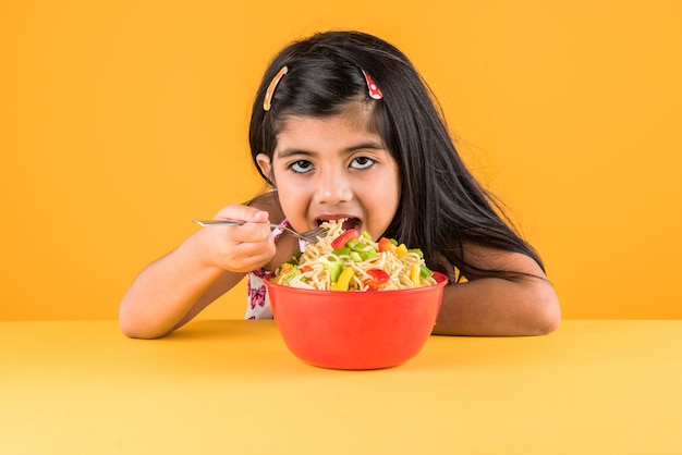 Linda niña india o asiática comiendo deliciosos fideos chinos con tenedor o palillos, aislado sobre fondo de colores