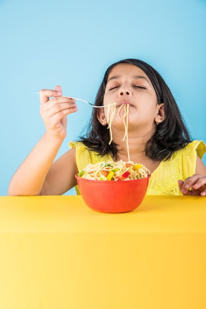 Foto linda niña india o asiática comiendo deliciosos fideos chinos con tenedor o palillos, aislado sobre fondo de colores