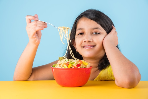 Linda niña india o asiática comiendo deliciosos fideos chinos con tenedor o palillos, aislado sobre fondo de colores