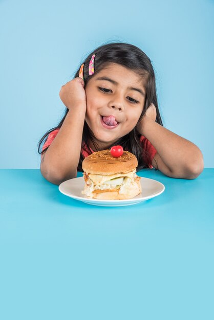 Foto linda niña india o asiática comiendo sabrosa hamburguesa, sándwich o pizza en un plato o caja. permanente aislado sobre fondo azul o amarillo.