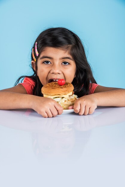 Foto linda niña india o asiática comiendo sabrosa hamburguesa, sándwich o pizza en un plato o caja. permanente aislado sobre fondo azul o amarillo.