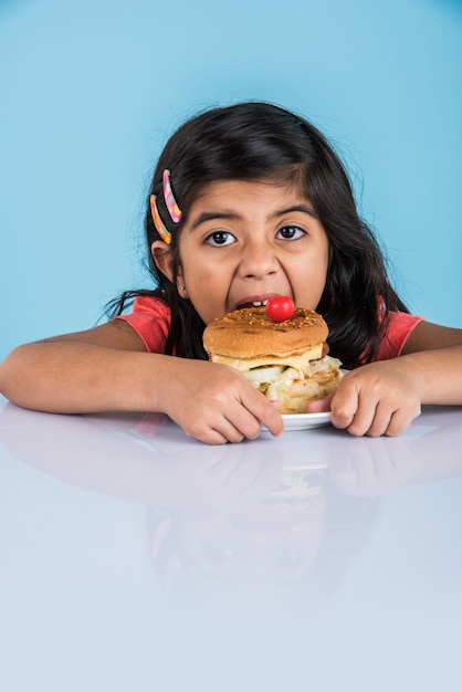 Linda niña india o asiática comiendo sabrosa hamburguesa, sándwich o pizza en un plato o caja. Permanente aislado sobre fondo azul o amarillo.