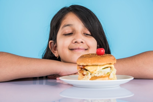 Linda niña india o asiática comiendo sabrosa hamburguesa, sándwich o pizza en un plato o caja. Permanente aislado sobre fondo azul o amarillo.