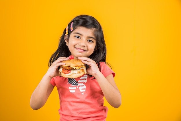 Linda niña india o asiática comiendo sabrosa hamburguesa, sándwich o pizza en un plato o caja. Permanente aislado sobre fondo azul o amarillo.