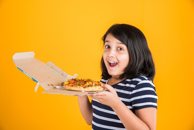 Linda niña india o asiática comiendo sabrosa hamburguesa, sándwich o pizza en un plato o caja. Permanente aislado sobre fondo azul o amarillo.