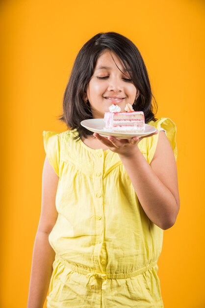 Linda niña india o asiática comiendo un trozo de pastel o pastel con sabor a fresa o chocolate en un plato. Aislado sobre fondo de colores