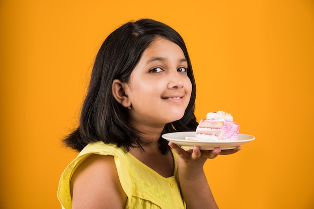 Linda niña india o asiática comiendo un trozo de pastel o pastel con sabor a fresa o chocolate en un plato. Aislado sobre fondo de colores