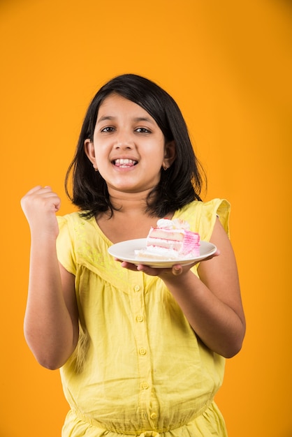 Linda niña india o asiática comiendo un trozo de pastel o pastel con sabor a fresa o chocolate en un plato. Aislado sobre fondo de colores