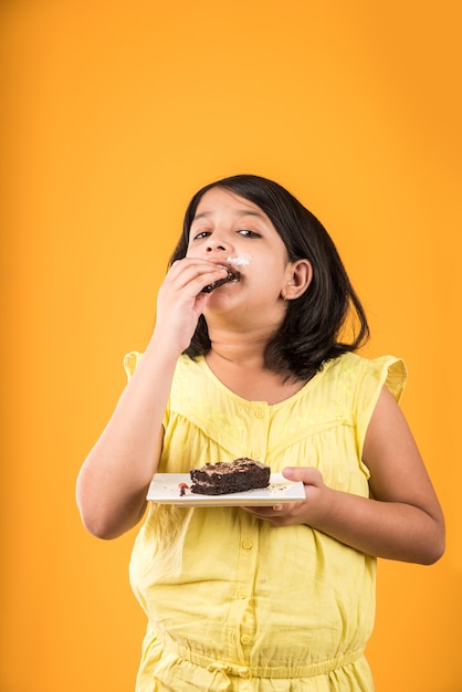 Linda niña india o asiática comiendo un trozo de pastel o pastel con sabor a fresa o chocolate en un plato. Aislado sobre fondo de colores