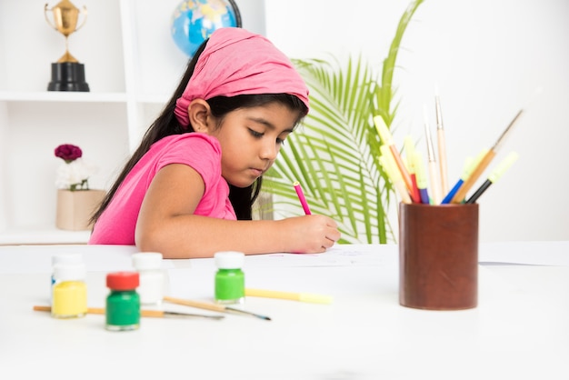 Linda niña india o asiática disfrutando de dibujar o pintar con pincel y pintura sobre papel en casa