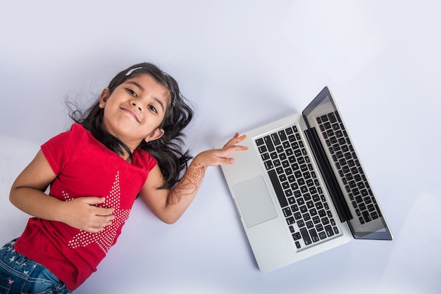 Foto linda niña india o asiática que estudia en la computadora portátil o trabaja en un proyecto escolar mientras está acostado o sentado en el suelo, aislado sobre fondo blanco.