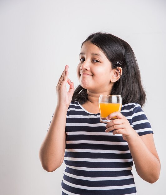 Linda niña juguetona india o asiática bebiendo mango fresco o jugo de naranja o bebida fría o bebida en un vaso, aislado sobre fondo blanco.