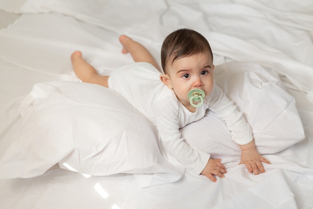 Linda niña mestiza acostada en la cama con un chupete en la boca con un traje blanco con ropa de cama de algodón. Foto de alta calidad