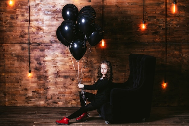 Linda niña de moda con un montón de globos negros contra la pared con lámparas