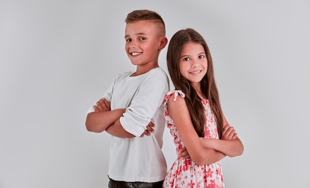 Linda niña y niño sobre un fondo gris están de espaldas, con los brazos cruzados y sonriendo.