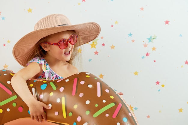 Linda niña niño con sombrero con anillo inflable en la pared de color. Cuarentena vacaciones de verano en casa