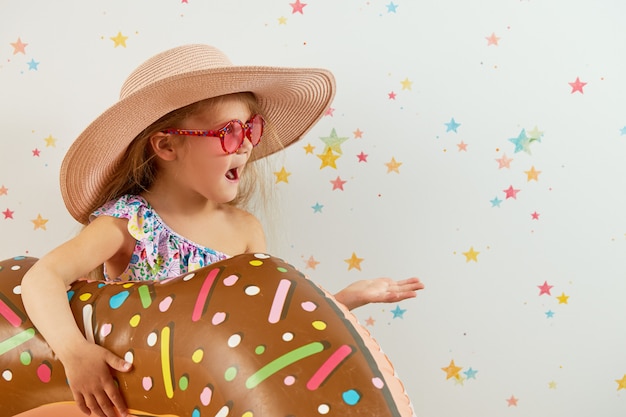 Linda niña niño con sombrero con anillo inflable en la pared de color. Cuarentena vacaciones de verano en casa