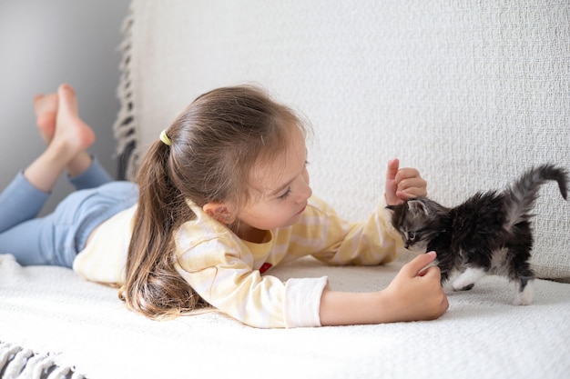 Una linda niña de pelo largo de 5 años se acuesta en el sofá y juega con un gatito de la raza Maine Coon Habitación brillante Amistad entre el hombre y el gato