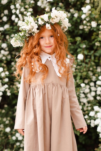 Una linda niña con el pelo rojo largo y rizado usa una corona floral y un vestido elegante sobre el fondo de la naturaleza