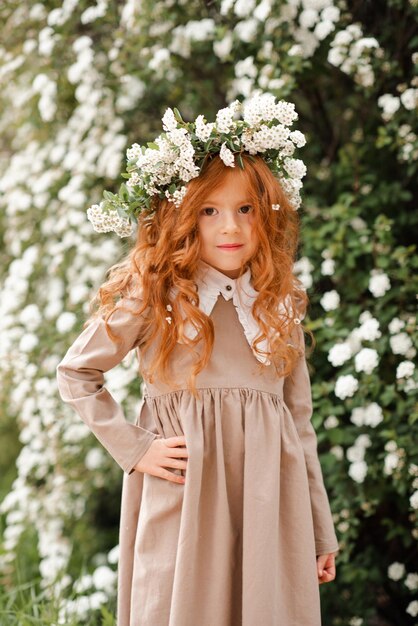 Una linda niña con el pelo rojo largo y rizado usa una corona floral y un vestido elegante sobre el fondo de la naturaleza