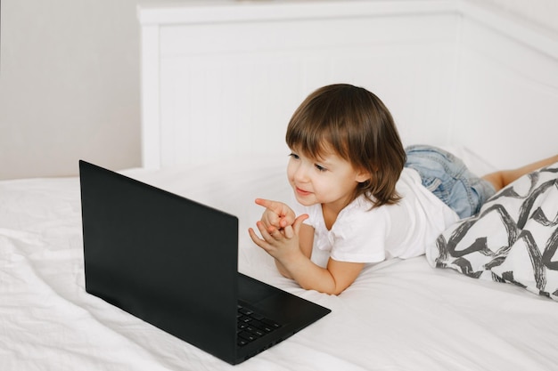 Una linda niña pequeña acostada en la cama usando una laptop