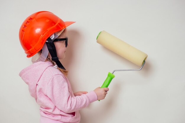 Linda y niña pequeña en un casco de construcción, casco y gafas con rodillo de pintura, pinta una pared blanca en casa, futuro diseñador, mujer de negocios, arquitecto, orientación profesional, concepto