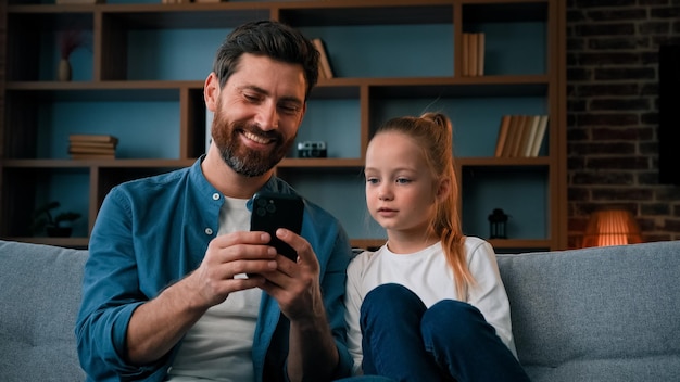 Linda niña pequeña descansando en un sofá acogedor con un joven padre cariñoso jugando en