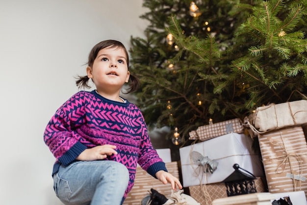 Una linda niña pequeña jugando con adornos bajo un árbol de Navidad decorado en el suelo de la habitación Feliz Navidad y Felices Fiestas