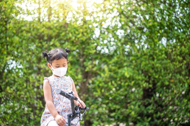 Linda niña pequeña con mascarilla protectora en bicicleta al aire libre