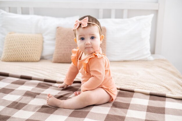 Una linda niña pequeña con un traje rosa está sentada en una cama con ropa de cama blanca con una manta beige en casa en el dormitorio mirando a la cámara sonriendo