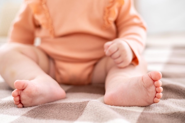 Foto una linda niña pequeña con un traje rosa está sentada en una cama con ropa de cama blanca con una tela escocesa beige en casa en el dormitorio, las piernas desnudas del bebé, los dedos de los pies del bebé